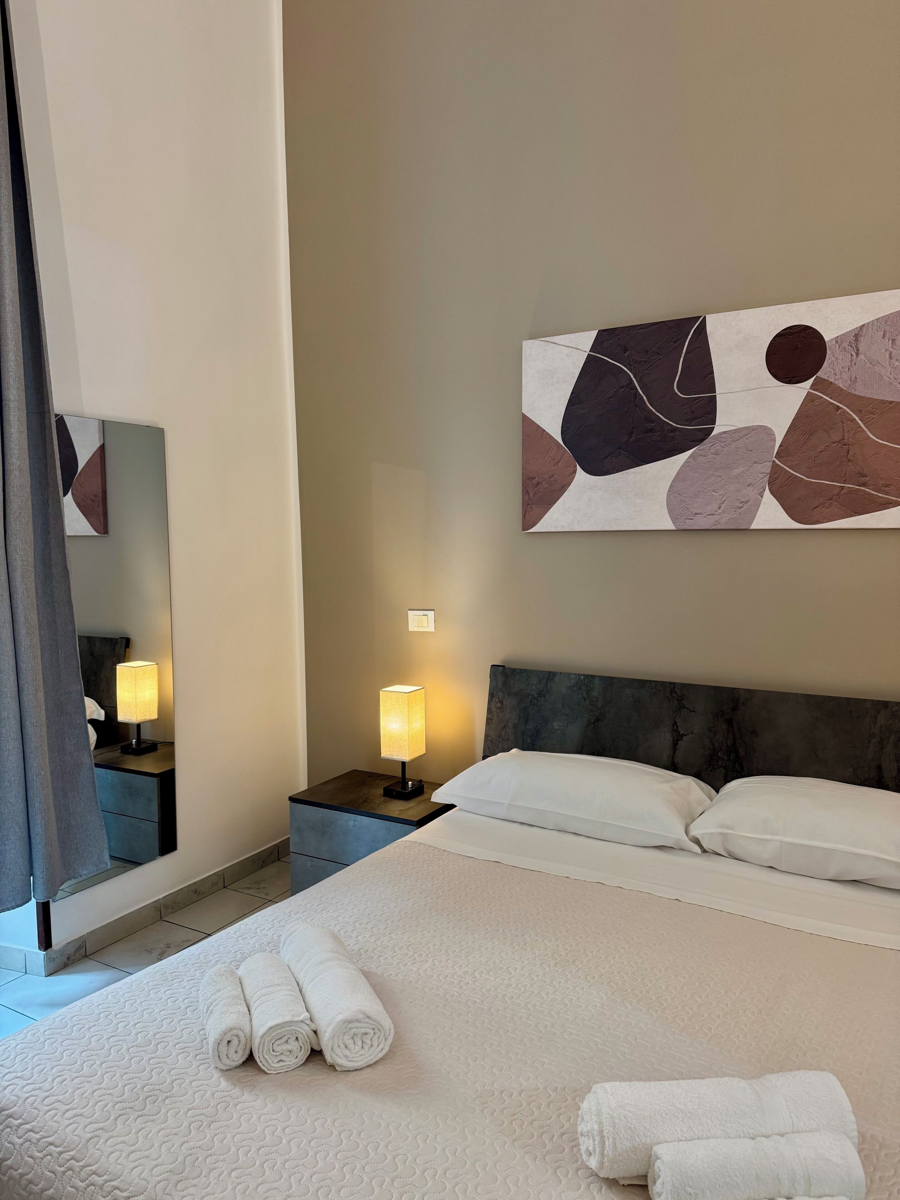 A hotel room with a bed, two bedside lamps, a large abstract painting, and a mirror.