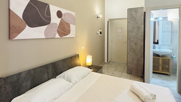 A hotel room with a bed, a nightstand, a wall-mounted lamp, a painting, and a bathroom visible through an open door. - Bosuites Rome (Rome)