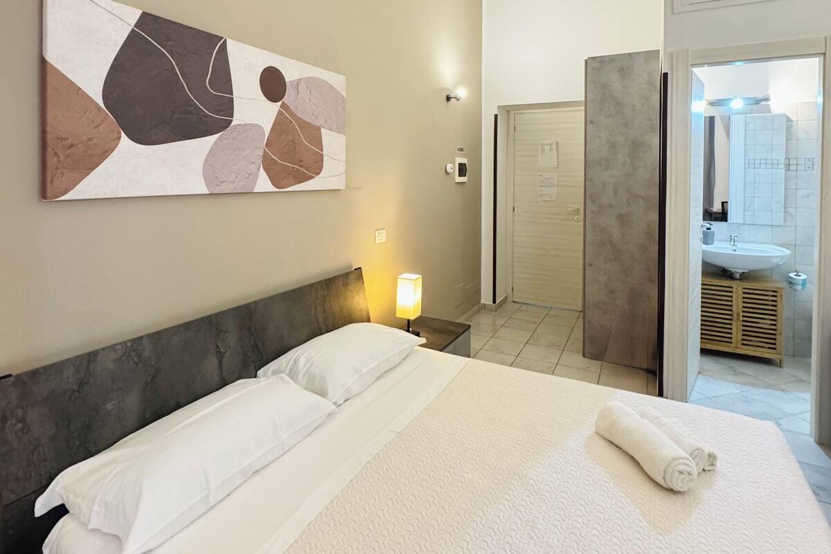 A hotel room with a bed, a nightstand, a wall-mounted lamp, a painting, and a bathroom visible through an open door.