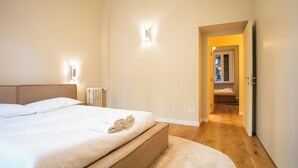 Ein Schlafzimmer mit Bett, Nachttischlampen, einem Heizkörper und Blick in einen anderen Raum durch eine offene Tür.