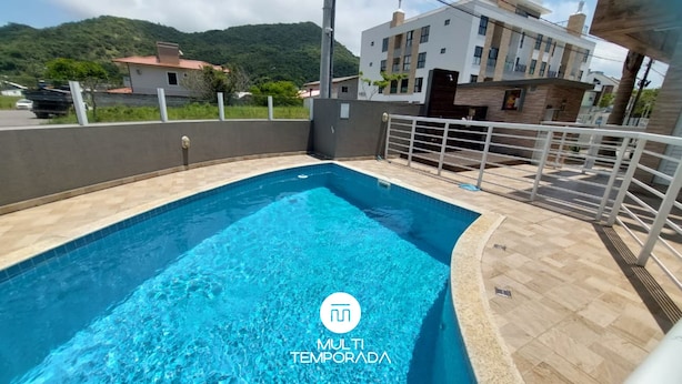 A swimming pool with a tiled edge, a metal fence, and a view of a multi-story building in the background. - Apartamento Aconchego do Ribeirão - FLN (Florianópolis)