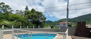 Piscina com deck branco e grade de metal, cercada por uma cerca, com vista para uma montanha e casas ao fundo.