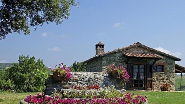 A stone house with a tiled roof, a chimney, and a flower bed with pink flowers. - Antico Casale San Lorenzo (Toscana)