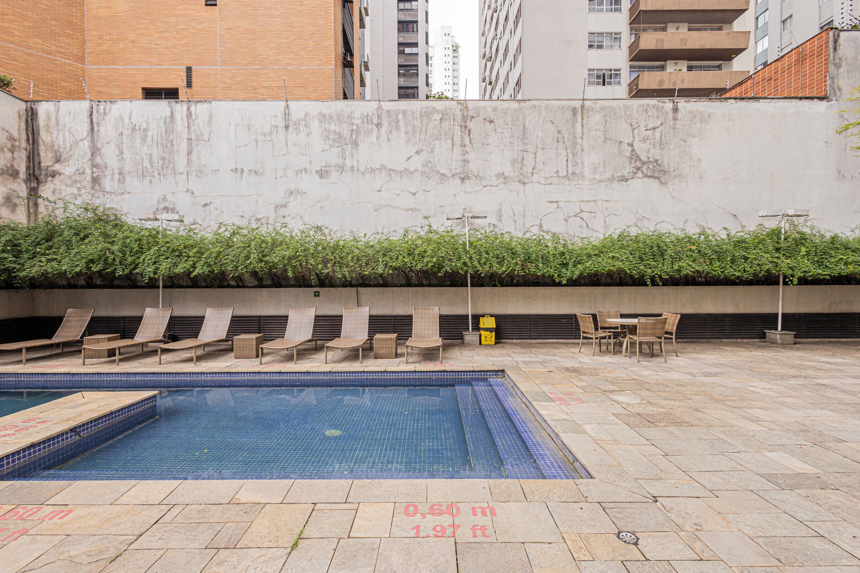A swimming pool with lounge chairs and a table, surrounded by a concrete wall and greenery.