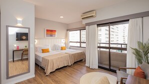 A hotel room with two beds, a desk, a chair, a TV, and large windows with a view of buildings. - QSP - Jardim Paulista by Anora Spaces (São Paulo)