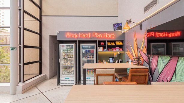 A modern interior with a vending machine, a neon sign reading 'Work Hard, Play Hard,' and a table with chairs. - QSP - Jardim Paulista by Anora Spaces (São Paulo)