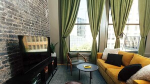 Un salon avec un mur en briques, un canapé jaune, un pouf en cuir marron et une petite table ronde.