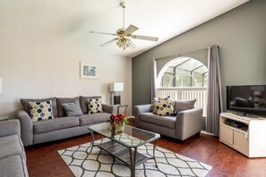 A living room with a sofa, armchair, glass coffee table, ceiling fan, TV, and a large window with curtains. - Enchanted 3BR pool home 10 mins to Disney (Kissimmee)