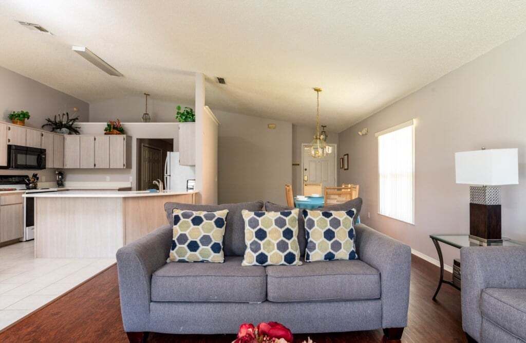 A modern living room with a grey sofa, patterned cushions, a dining area, and a kitchen in the background.