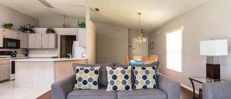 Ein modernes Wohnzimmer mit grauem Sofa, gemusterten Kissen, einem Essbereich und einer Küche im Hintergrund.