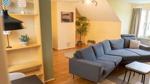 A modern living room with a grey sectional sofa, a dining table with chairs, a small kitchen area, and a ceiling fan. - Harbor-View Loft | 2BR Near City (Gothenburg)