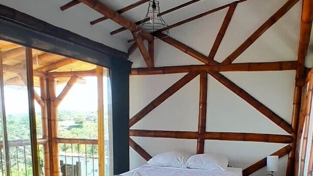 A bed with a white bedspread and pillows, a nightstand with a lamp, and a view of greenery through large windows. - Cozy Lodging in the coffee growing region (Ulloa)