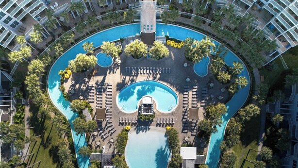 Una vista aerea di un resort con una piscina centrale, diverse aree relax e palme circostanti.