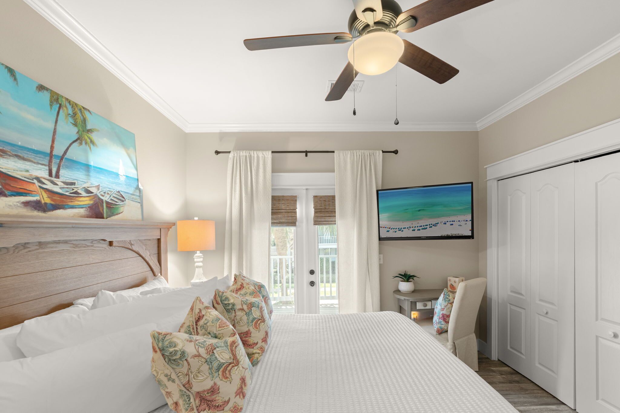 A bedroom with a bed, a ceiling fan, a TV, a painting, and a view through a door.