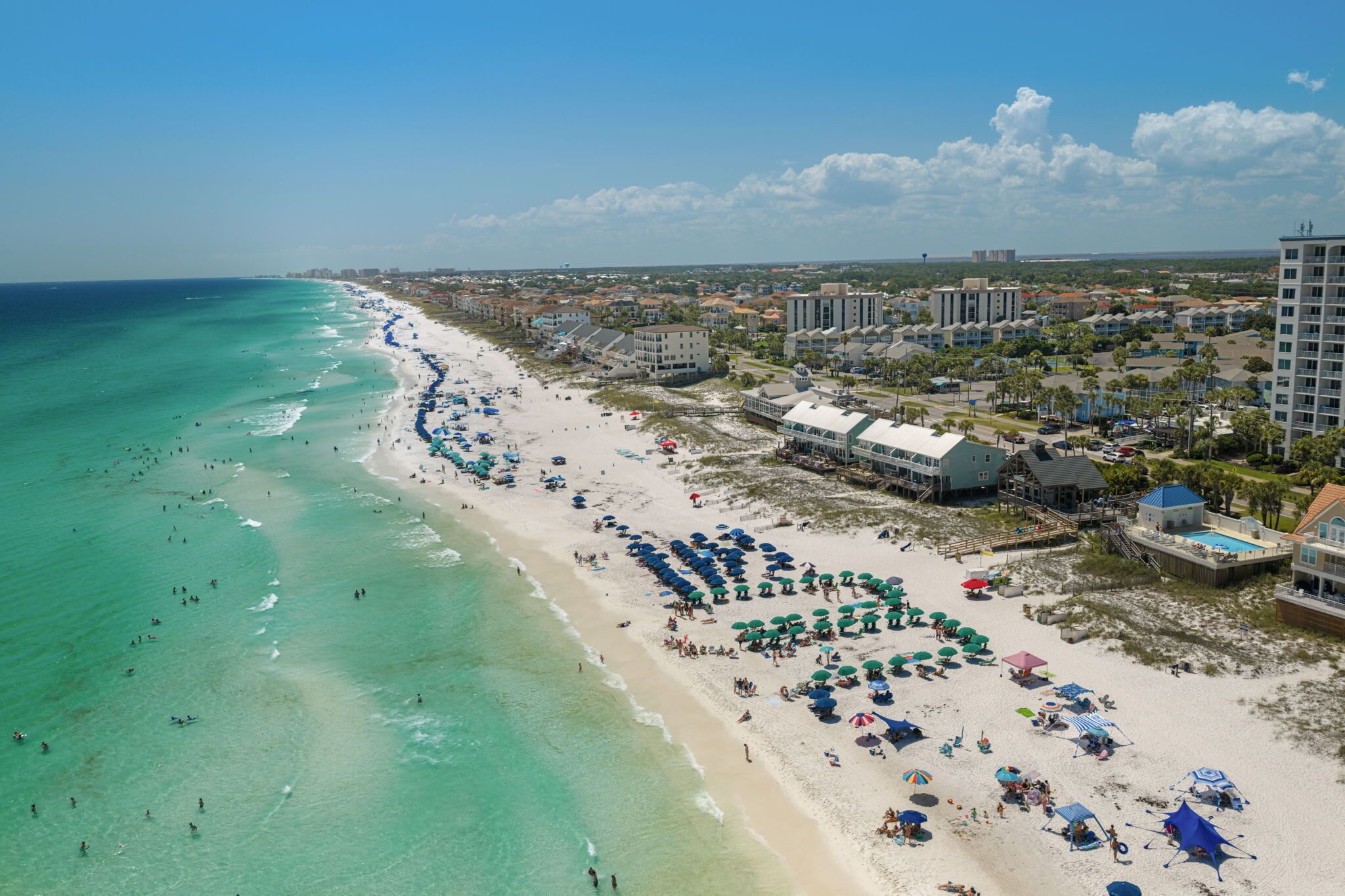A beach resort with a long sandy shore, multiple beach umbrellas, and a mix of residential and commercial buildings.
