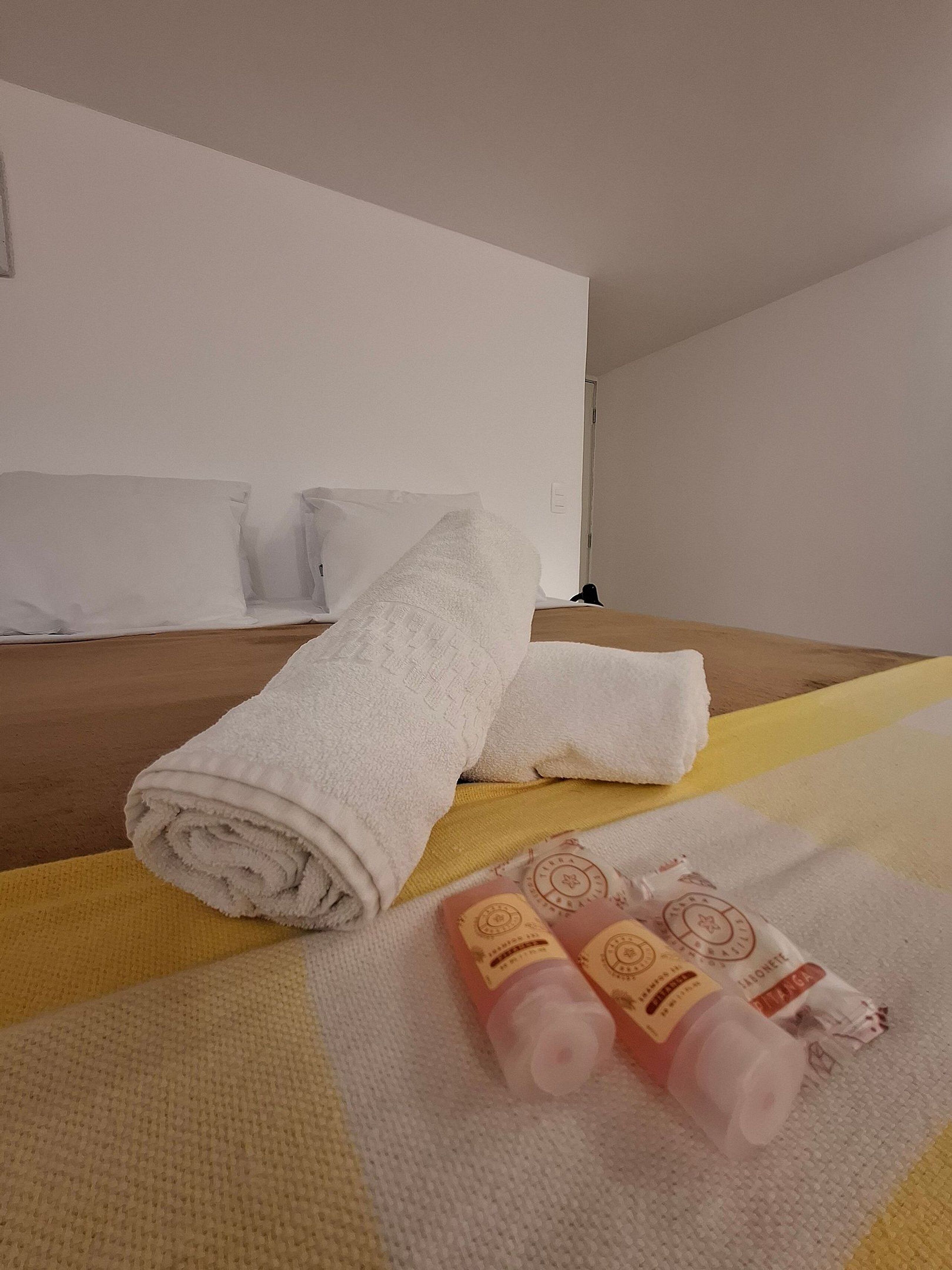 Ein ordentlich bezogenes Bett mit aufgerollten weißen Handtüchern und zwei Lotionflaschen auf einer gelb-weißen Tagesdecke mit Struktur.