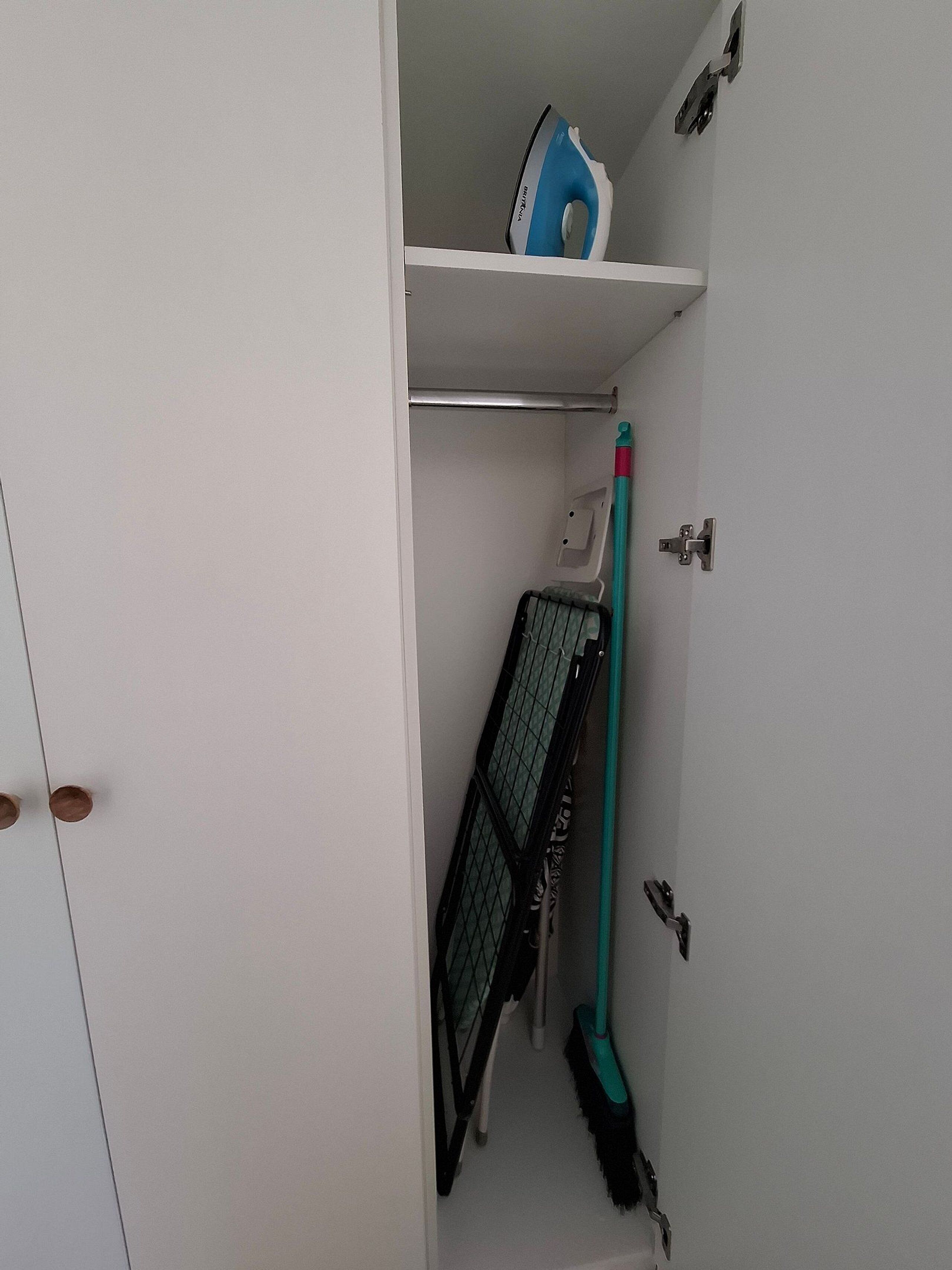 A white wardrobe with a hanging rack, a blue iron, and a broom.