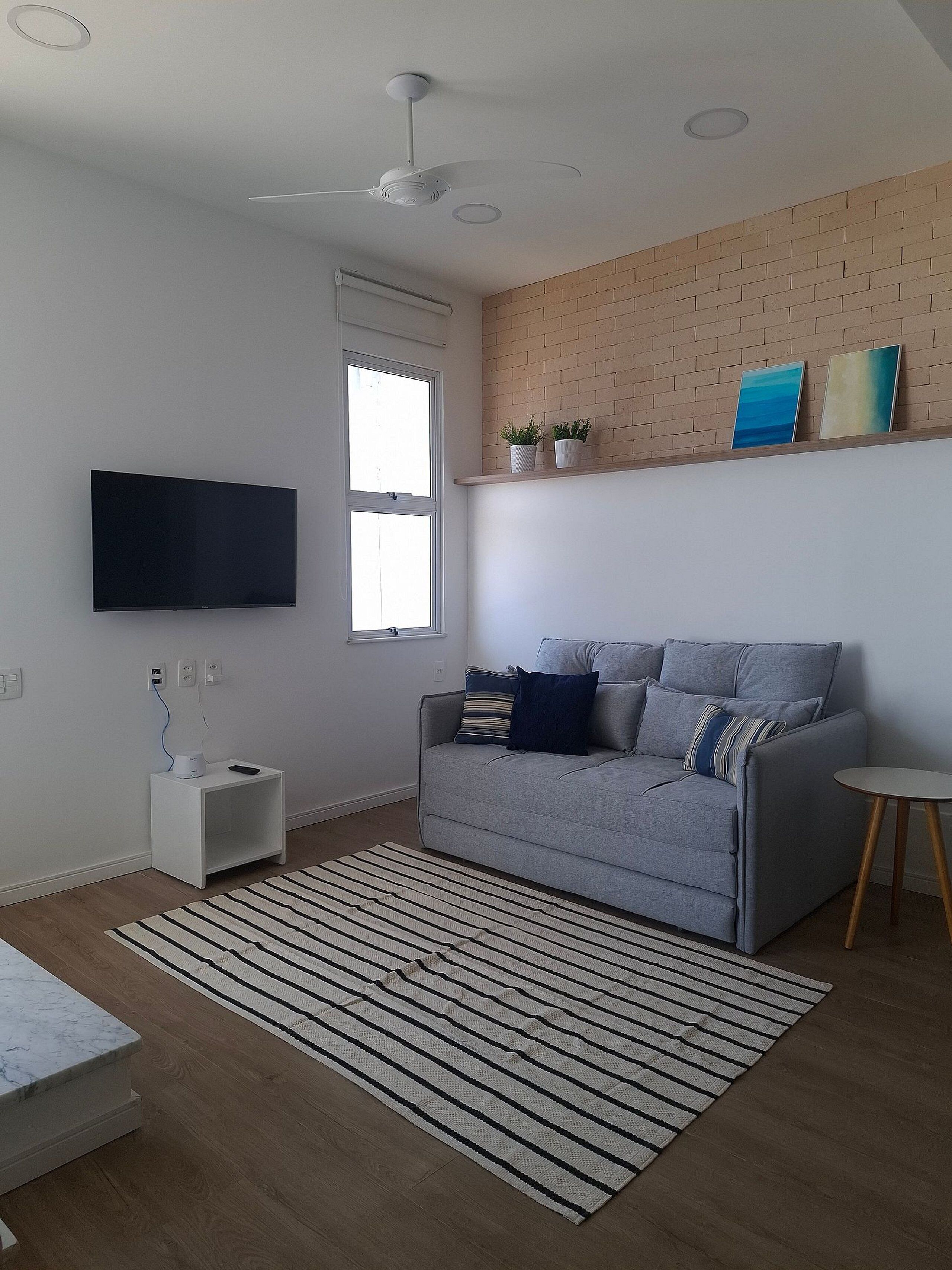 A modern living room with a grey sofa, a flat-screen TV, a ceiling fan, a window, and a brick wall.