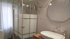 Ein Badezimmer mit Duschkabine aus Glas, einem Holztischchen mit weißem Waschbecken und einer Wand mit Marmorfliesen.