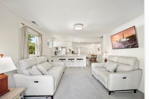 Ein modernes Wohnzimmer mit zwei weißen Ledersofas, einem Küchenbereich im Hintergrund und einem großen Flachbildfernseher an der Wand.