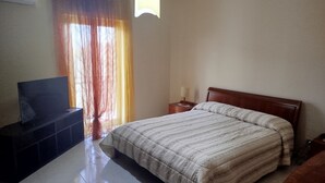 Un dormitorio con una cama, un televisor sobre un mueble, una mesita de noche y una ventana con cortinas.