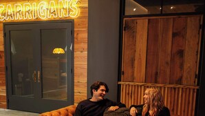 Un coin salon accueillant avec un canapé en cuir et une table, un mur en bois orné d’un néon, et une grande porte en bois.
