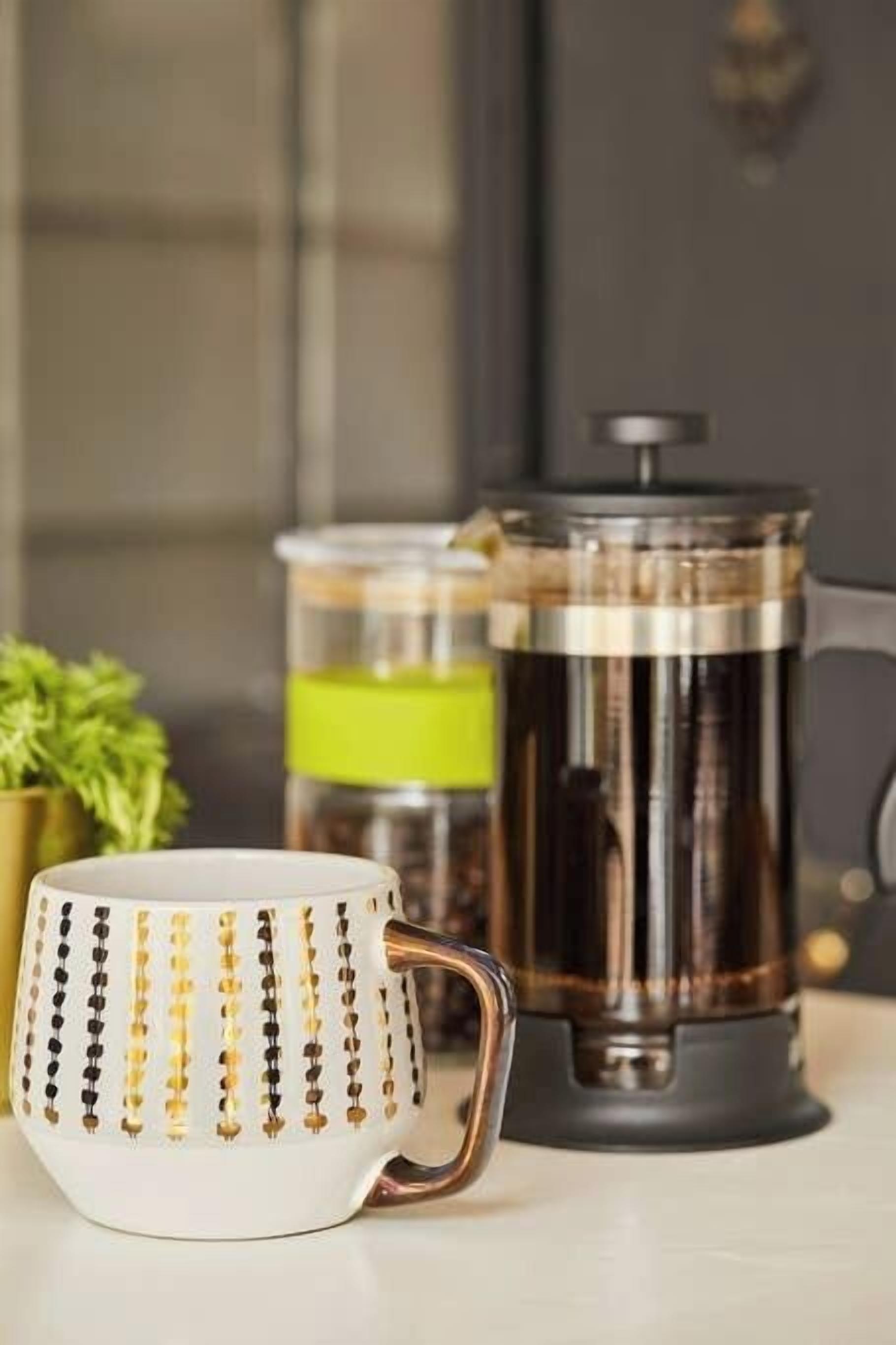 En vit mugg med guldfärgade pärlmönster och en metallisk kaffebryggare på en köksbänk.