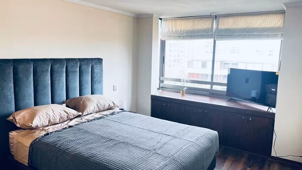 A bedroom with a bed, a window with curtains, a television on a wooden desk, and a small plant. - Providencia 2BR 2BA Apt + Private Parking (Providencia)