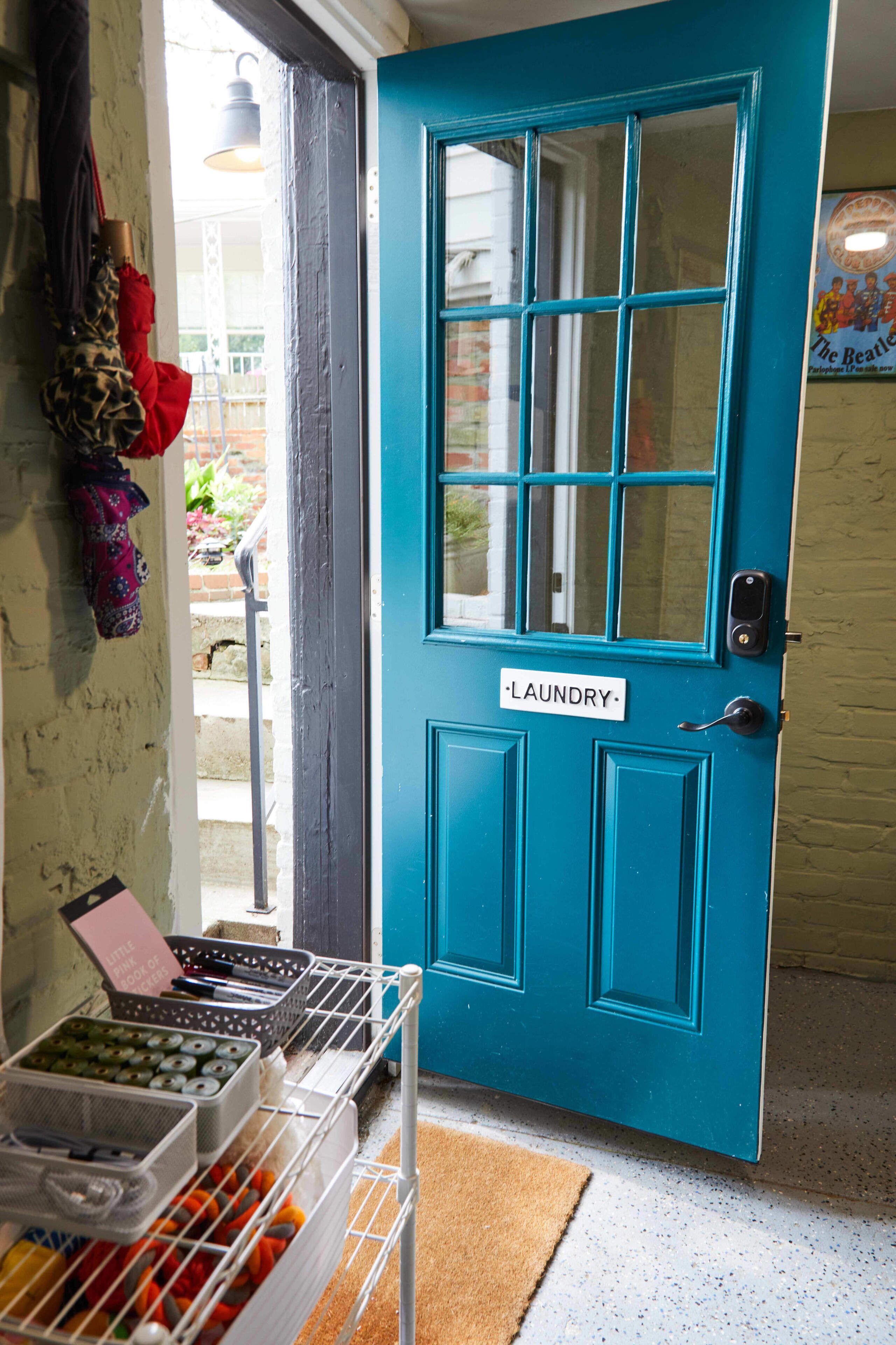 Une porte de couleur turquoise portant l’inscription « Buanderie », avec un étendoir en métal sur lequel sont posés des paniers à linge, et une petite table sur laquelle se trouve un objet rose.