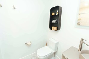 Un baño con inodoro blanco, lavamanos y estante con artículos de aseo.