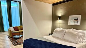 Hotellihuone, jossa on sänky, tuoli, pieni pöytä ja seinään kiinnitetty valaisin.