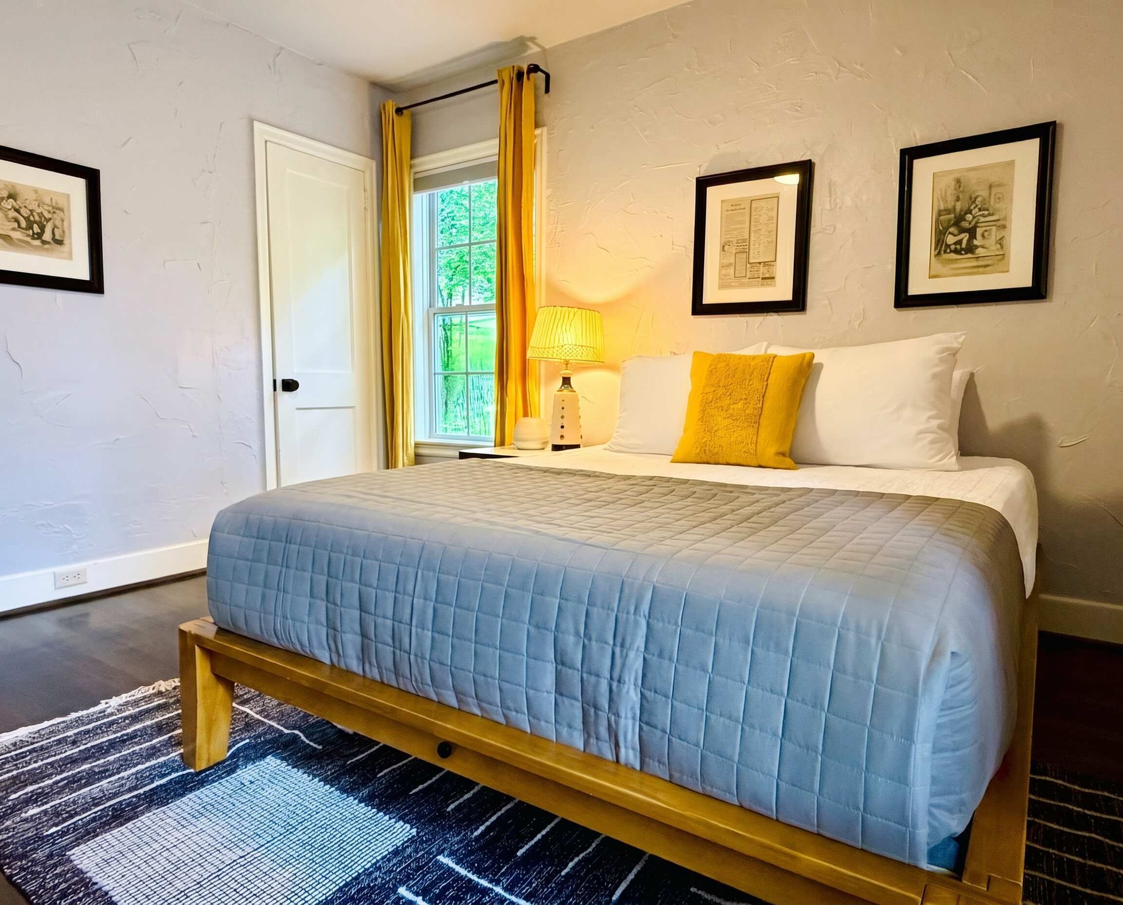 Ein Schlafzimmer mit Bett, einem Fenster mit Vorhängen, einer Lampe und gerahmten Bildern an der Wand.
