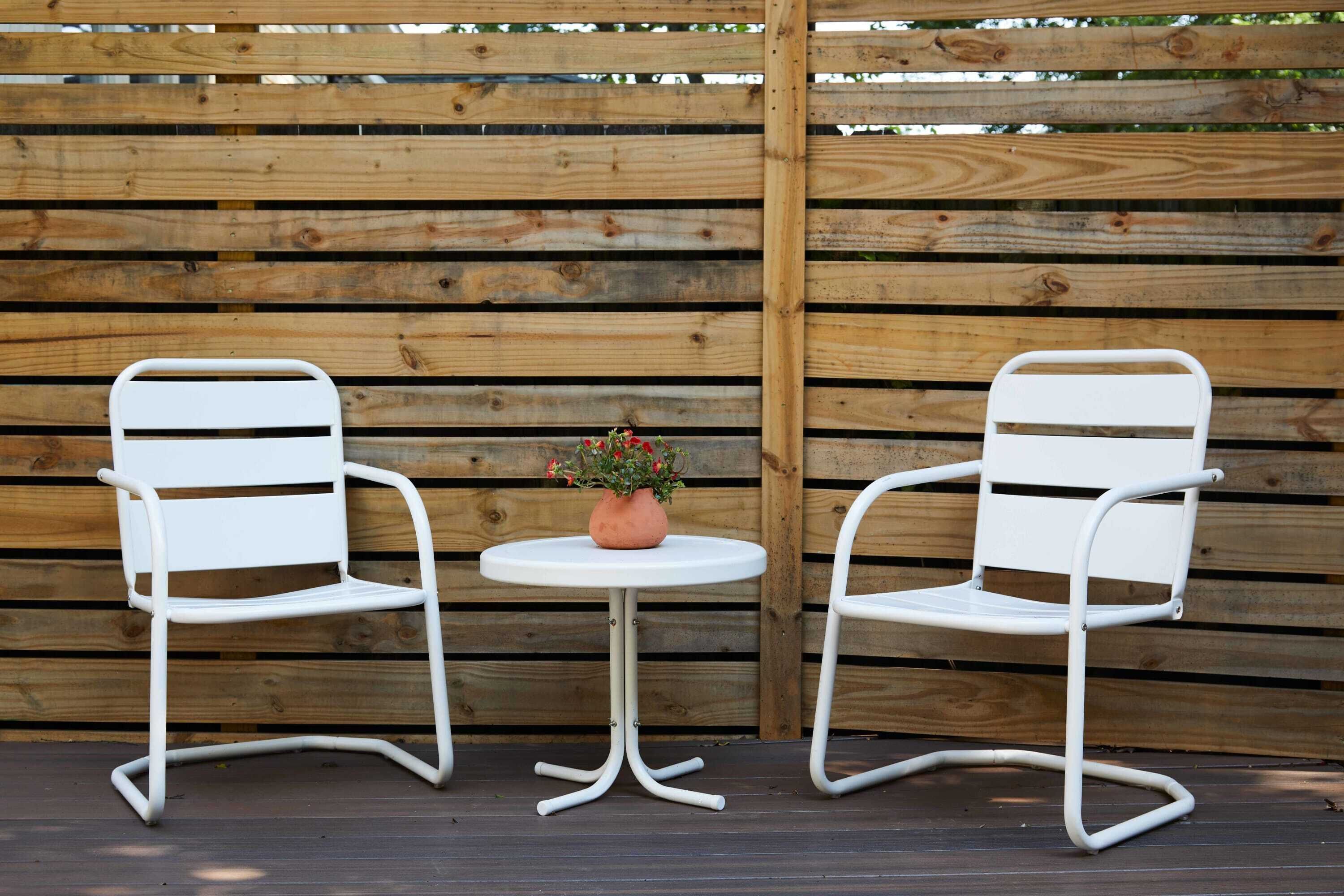Two white metal chairs with a small round table and a potted plant between them, set against a wooden fence background.