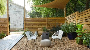 Eine Holzterrasse mit zwei weißen Stühlen und einem runden Hocker, umgeben von Kieselsteinen und Topfpflanzen.