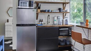 Eine kompakte Küche mit einem Edelstahl-Kühlschrank, einer Mikrowelle und einem Spülbecken. Über dem Spülbecken befindet sich ein Holzregal und ein Fenster mit Blick auf Grünflächen.