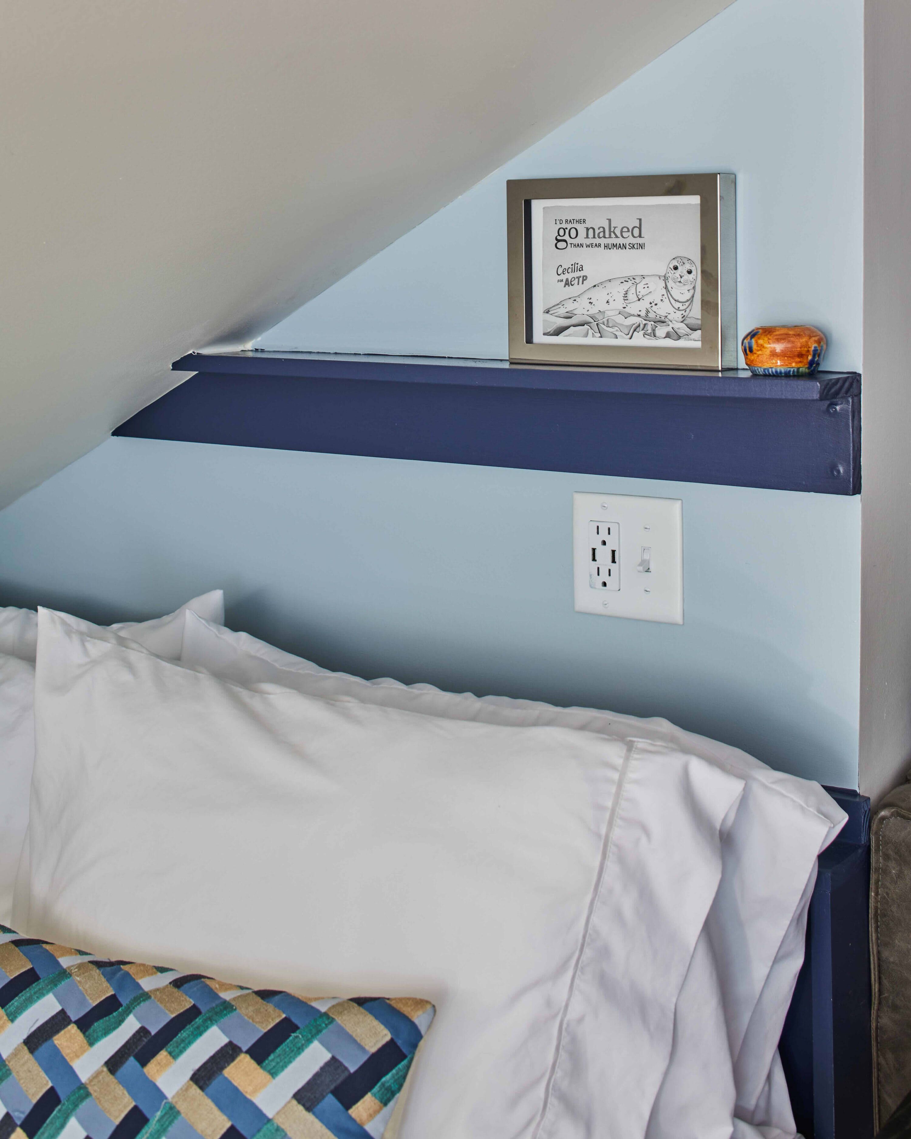 A bedroom with a bed, a pillow, a framed picture on a shelf, and a power outlet.