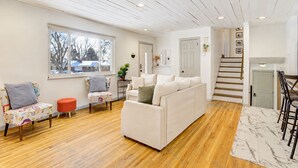 Ein Wohnzimmer mit weißem Sofa, einem bunten Sessel, einem roten Hocker, Holzboden und einem Teppich mit Marmoroptik. Eine Treppe führt zu einer zweiten Etage.