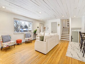 Un salon avec un canapé blanc, un fauteuil coloré, un pouf rouge, un plancher en bois, un tapis à motifs de marbre et un escalier menant à un deuxième étage.
