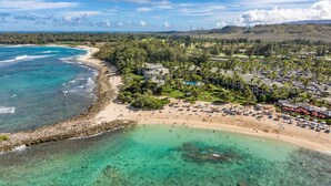 An aerial view of a coastal resort with a beach, clear waters, and a backdrop of hills and wind turbines.