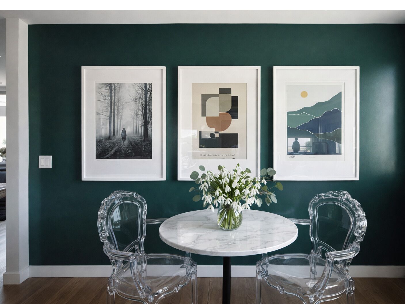 Un coin repas avec une table ronde, deux chaises et trois œuvres d’art encadrées sur un mur vert foncé.