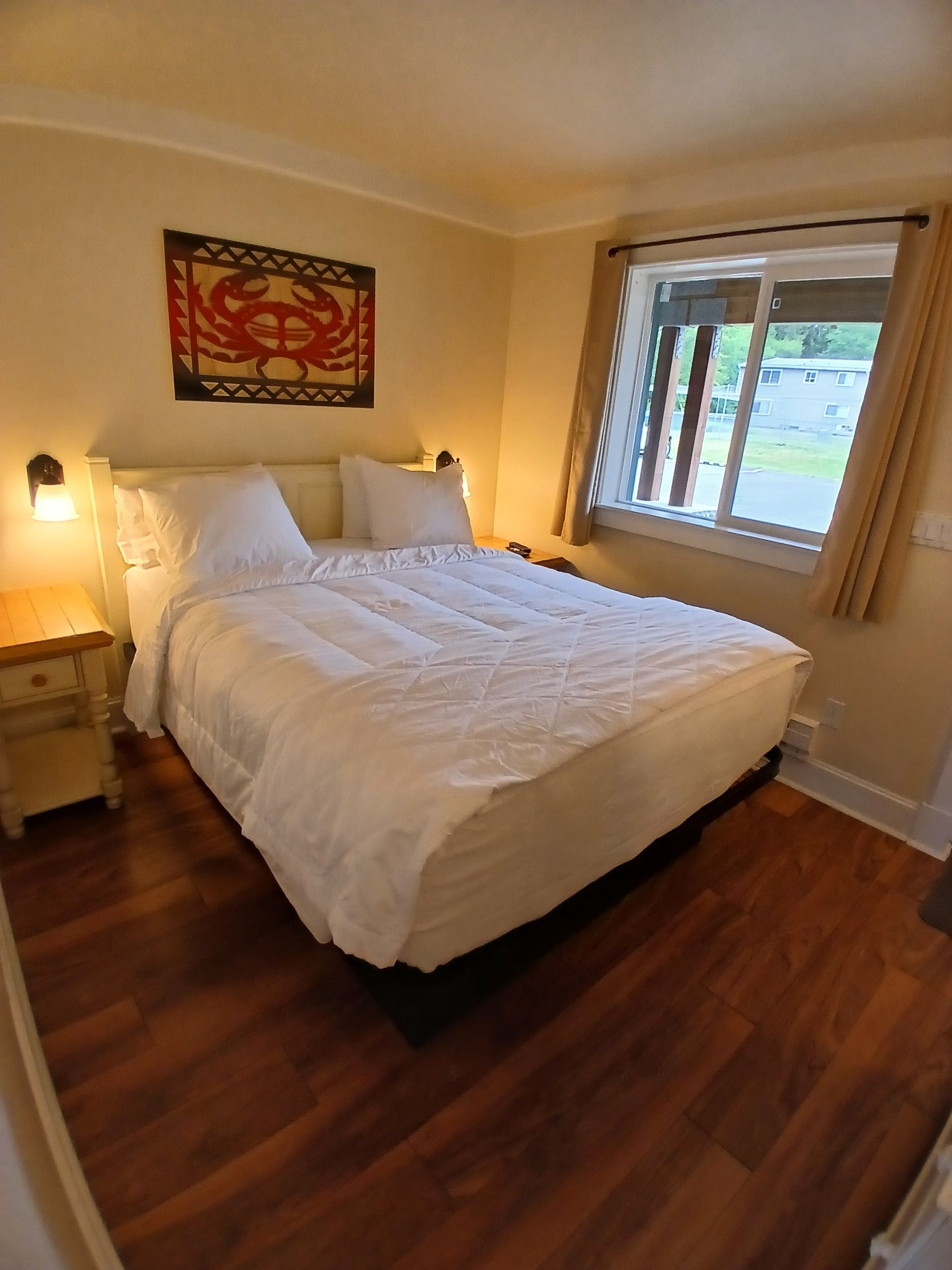 A neatly made bed with white linens in a room with wooden flooring, a window with curtains, and a wall-mounted artwork.