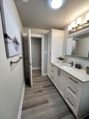 Une salle de bain moderne avec un lavabo blanc, un miroir et un meuble, une porte donnant sur un couloir, et un tableau encadré au mur.