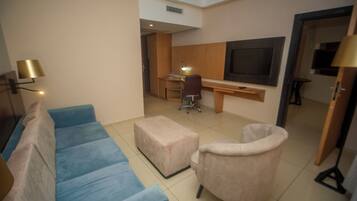 Habitación de hotel con un sofá azul, un pequeño taburete, una silla, un escritorio con su silla y un televisor montado en la pared.