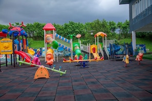 Une aire de jeux dotée de diverses installations, notamment des toboggans et des balançoires, aménagée sur une surface pavée, avec des arbres en arrière-plan.