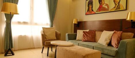 A hotel room with a sofa, armchair, and ottoman, a painting on the wall, and a window with curtains.