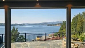 Une vue à travers de grandes portes vitrées donne sur un patio en pierre qui mène à une terrasse surplombant un lac, avec des montagnes qui se dessinent à l’horizon.