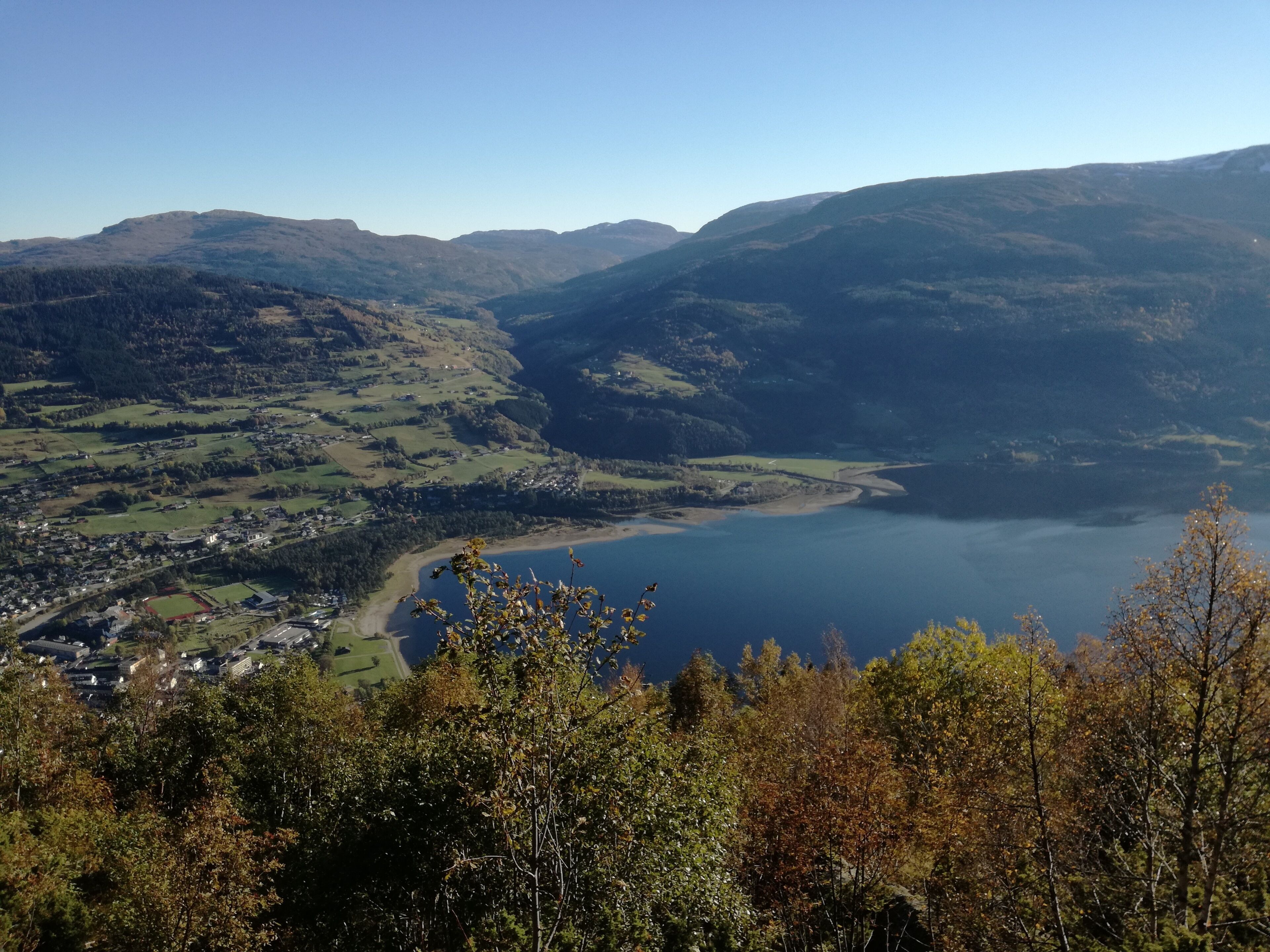 A panoramic view of a valley with a lake, surrounded by hills and trees.