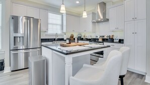 A modern kitchen with white cabinetry, stainless steel appliances, pendant lighting, and a central island with seating. - Masters/ANWA Rental Retreat (Grovetown)