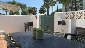 Un patio al aire libre con una mesa de madera, sillas, plantas en macetas y una pared decorativa.