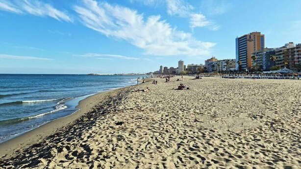 Una amplia playa de arena con un mar tranquilo, cielo despejado y edificios al fondo.